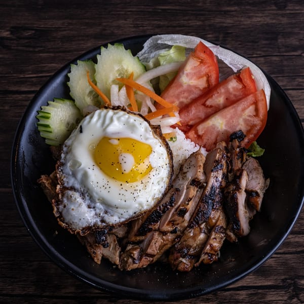 Grilled chicken, fried egg, rice, and fresh vegetables in a black bowl on a wooden table.