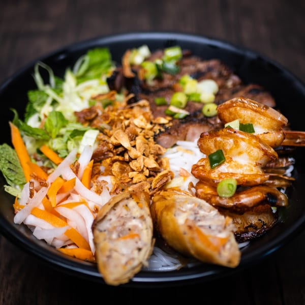 A black bowl of vermicelli noodles with grilled shrimp, pork, spring rolls, and vegetables.