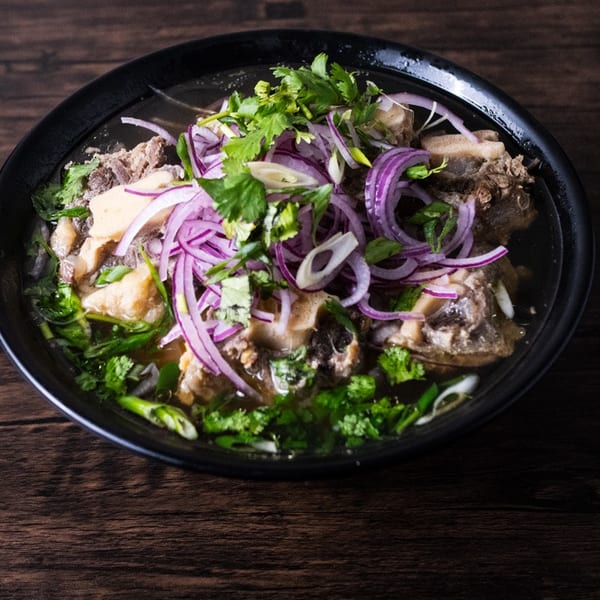 A black bowl of oxtail pho with red onions and cilantro on a dark wooden table.