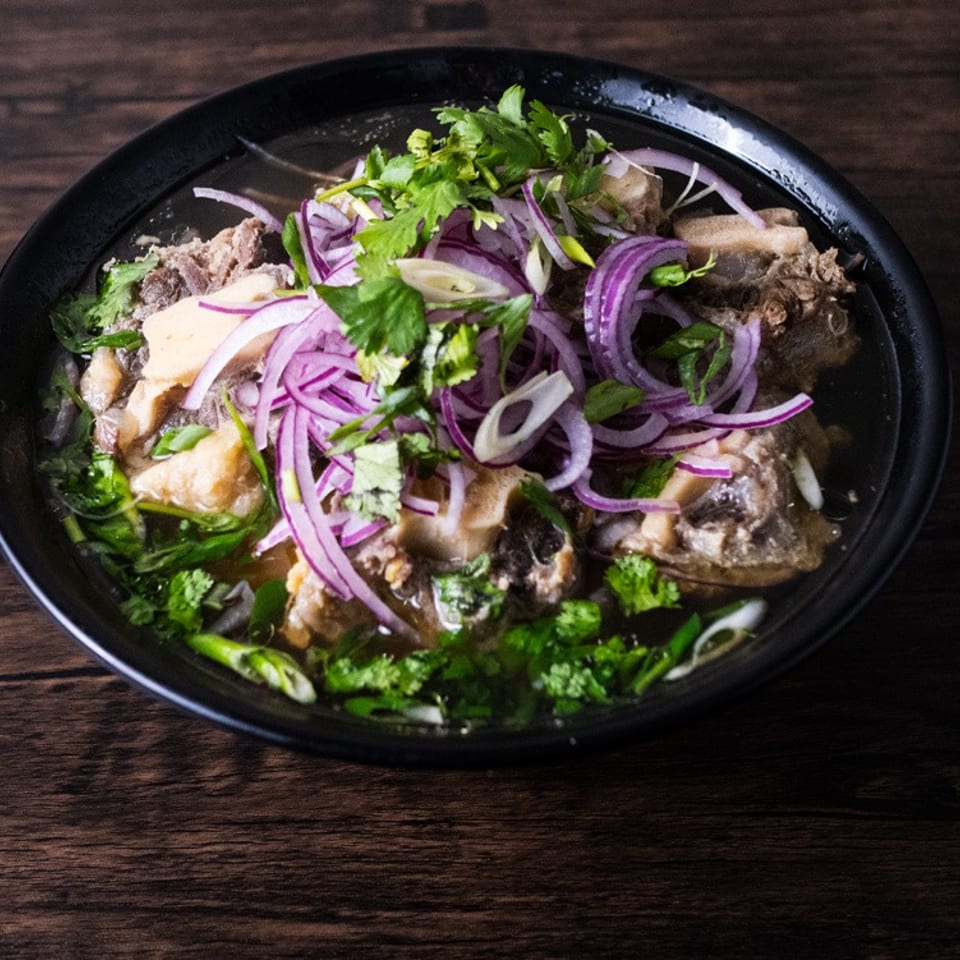 A black bowl of oxtail pho with red onions and cilantro on a dark wooden table.