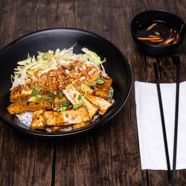 A black bowl of vermicelli noodles with tofu, vegetables, and sauce, with chopsticks and dipping sauce.